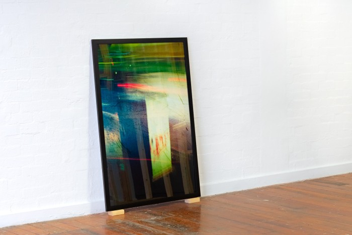 A framed photo of a bus driving past an elecitity box resting on two wooden blocks on the floor