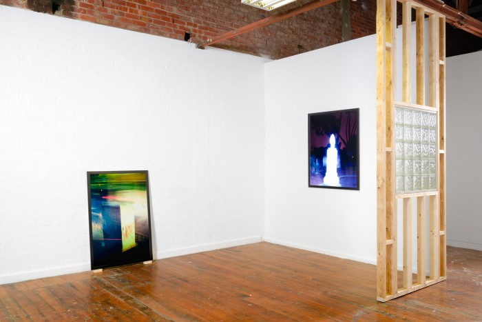 Installation view of a framed photo of a bus driving past an elecitity box resting on two wooden blocks on the floor, a framed photo of a glowing tombstone in a graveyard, and the wooden frame of a wall with a glass brick window in it.
