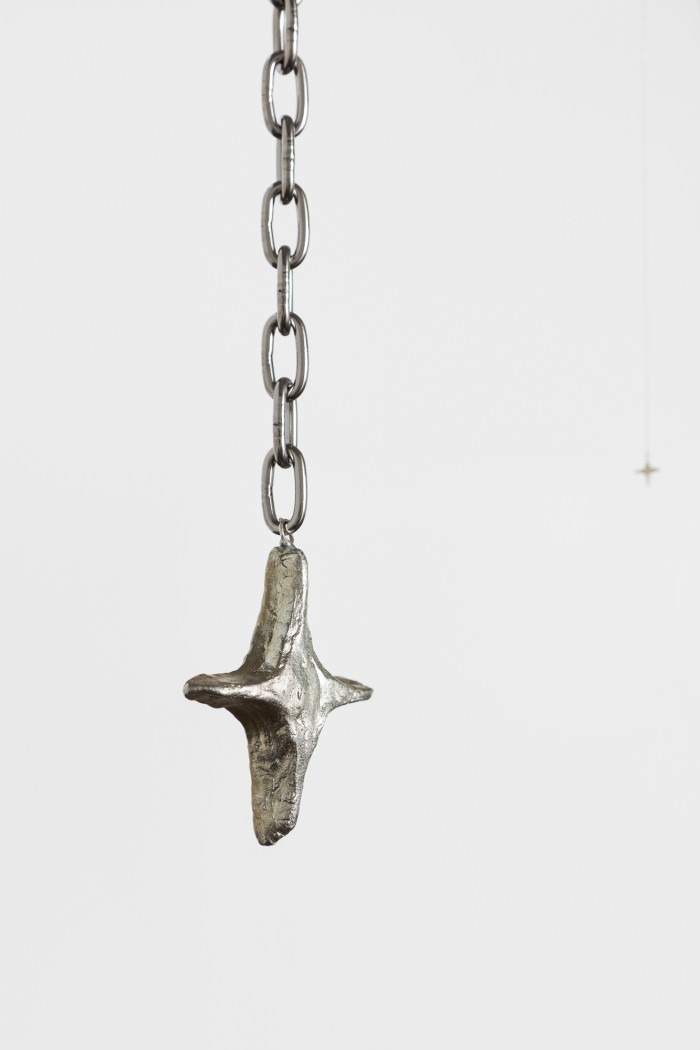 A four-pointed star scultpure, sliver in colour, suspended from a lightweight chain on a white background. There is another star in the distance, out of focus.