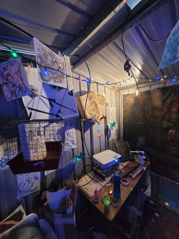 Image of Installation inside of shed, there is a table in the corner with a projector on it as well as small pieces of wood. There is a light that shines on a wishbone hung from the wall and an iron rectangle, around 1.5 metres high, with a memory foam pillow shoved on it with a playful smile and two dish glove hands out as if to wave, holding a tattslotto ticket in one arm. There is a cage with a canvas inside it with diary clippings and dog drawings and a picture of a possum in a photograph.