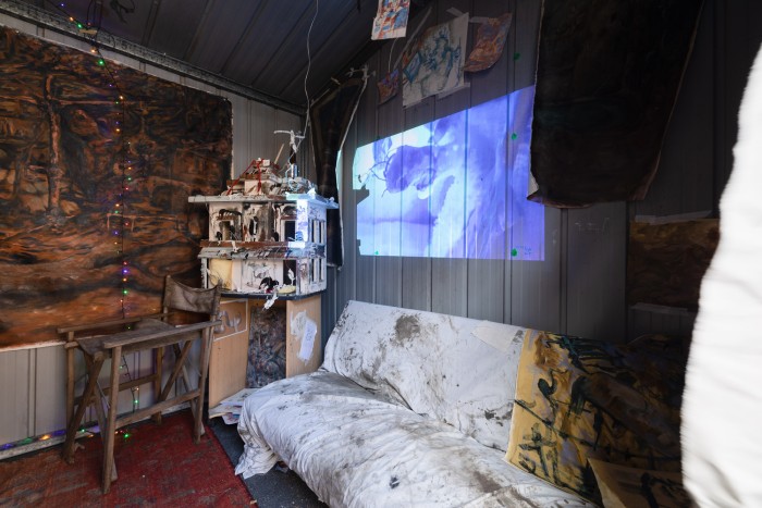 Image of installation inside of shed, a couch with a painted dirty sheet on it, a projected image of a cartoonish face above it (the still of the video work), a dolls house on a small table with small sculptures of humans and animals with clay and found objects. Various small works slightly out of shot hung by pegs.
