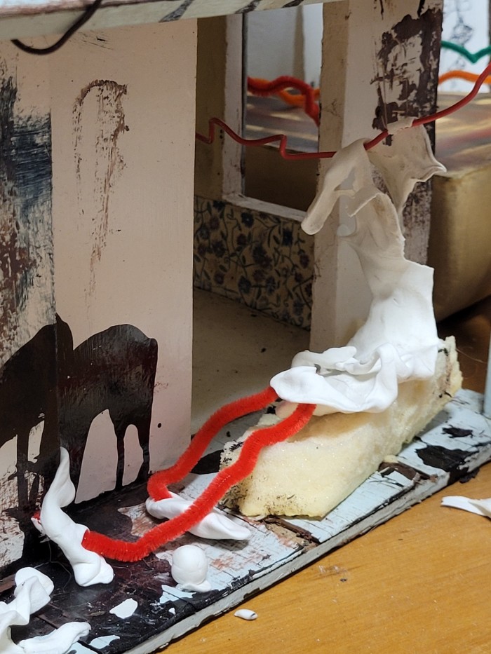 Close up of human figure playing fetch, small pipecleaner legs in a recline under the roof of the dollhouse on a piece of memory foam with the top of the body wrapped around a piece of wire.