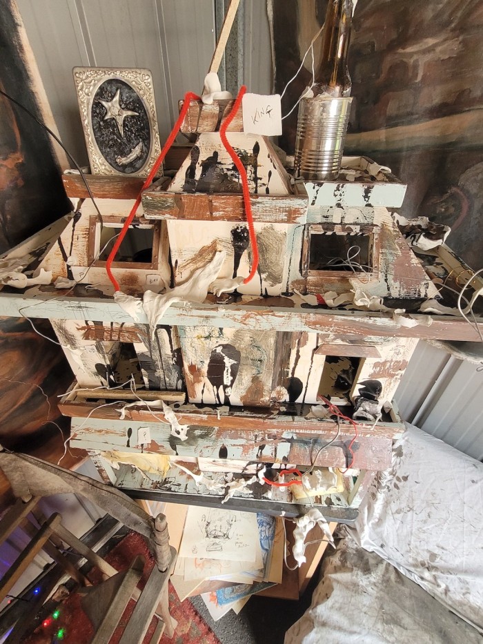 Dollhouse with animals and humans that are depicted as melted and abstract, small and dotted around the building. There is a label at the top that says "king" with a figure with long pipecleaner legs and a mouth open in a slight grin and one below that says "playing fetch?" with a melted human-like figure and a stuck dog. There is a frame on top with a dried lizard lying under a star made out of clay with bits of gravel around it and a beer bottle with clay on top of it in a cross-like shape.