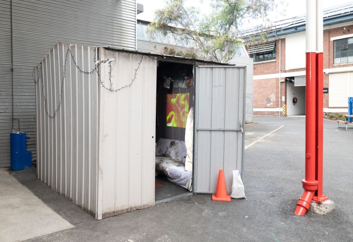 Picture of a small shed (2.5m x 2.5m) outside under the VCA building with christmas lights drapped around the roof gently, the door is open with a traffic cone outside and a video and couch visible within.