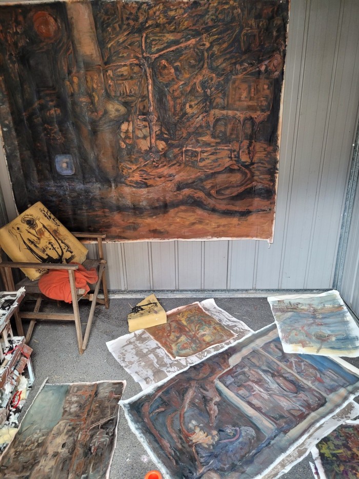 Paintings on the ground of a shed on loose canvas sitting like rugs, depicting abstract oil painted cities, a painted city on the wall with a hillshoist, dog and person in the middle melting into the ground, a dollhouse in the corner and a chair with a memory foam pillow sitting on it with a smiley face painted on with ink.