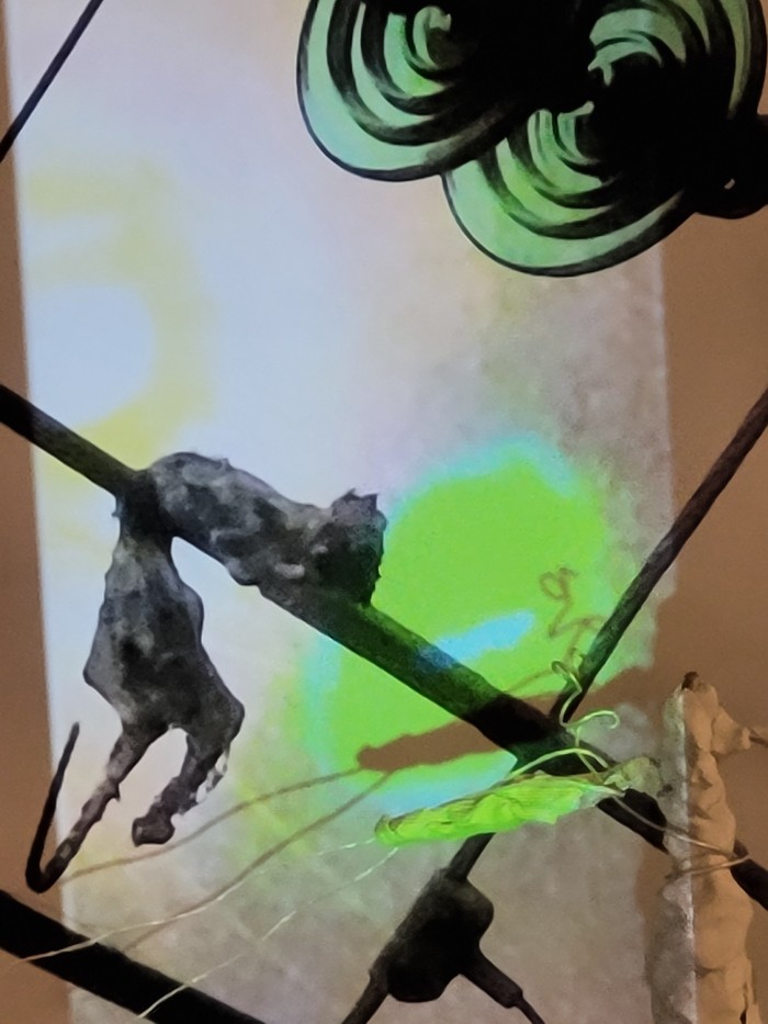 A possum on a powerline that is fried and dead, there is only sky and a plastic cone part of the connecting powerline visible. There is a cross-like sculpture infront of the image with a green light over both the possum and sculpture.