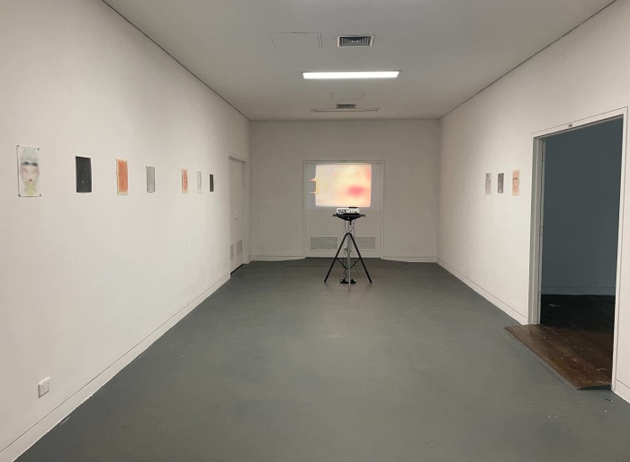 View of the room from the point of entry, slide projector stand, projector and super 8 film is central, and the film is projected onto two doors at the back of the room. Drawings are hung on the walls on eitheir side of this and are colour and black and white, and the super 8 film is in colour. The lights are on in the room except for a row that is off above the projection.