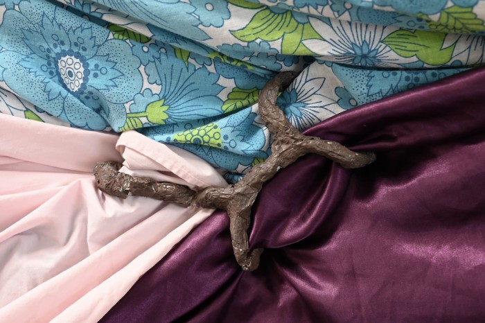 This is a detailed shot of the small sculptures which help suspend the fabric on the cage wall. A light brown organic tree branch style sculpture is in the centre of the frame holding together three different coloured fabrics.