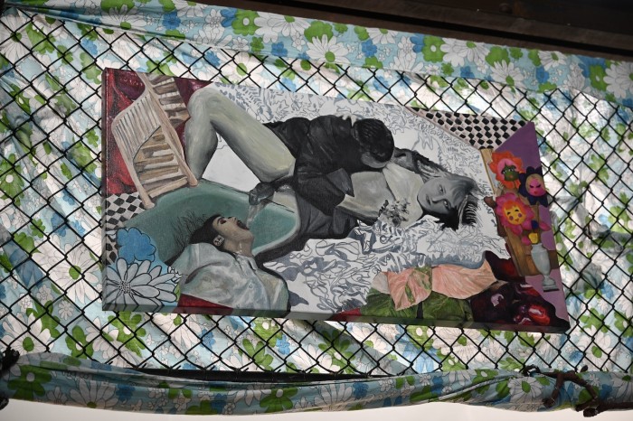 There is a horizontal painting suspended on a black wire cage wall. Behind the painting and the metal cage its suspended on is floral bedsheet fabric. The painting itself is a collage of images that have been repainted in a realistic style. The focus of the painting is a woman lying down looking at the camera while a masculine figure lift her shirt and nuzzles her breast. Extending from the female figure's pubic mound is a bathtub tap. There is water flowing out of the tap and into another female figure who's lying underneath in a teal coloured bathtub. Surrounding these figures is a spiral staircase, black and white kitchen tiles, monstera plant leafs. Other collaged figures in the painting is an earn with three cartoon style flowers coming out of it and a child’s hand reaching around behind a door to touch it. In the gaps of the painting are large pomegranate seeds that have also been collaged and painted.