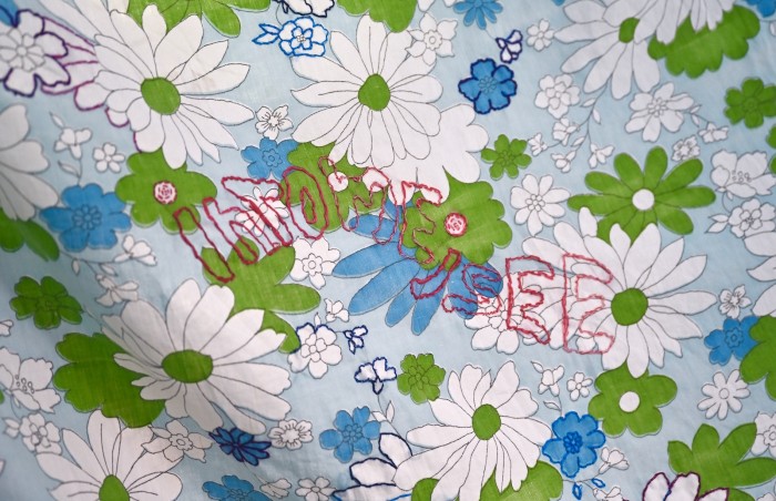 On floral vintage bedsheet there is a red cotton thread embroidery which reads in bubble style text 'intomeisee'. Some of the flowers on the bedsheet are outlined in blue and purple embroidery threads