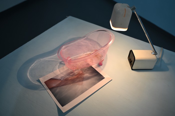 Top down view of large square plinth painted pale blue. There is a vintage desk lamp pointing down on a pale pink minature bathtub. Underneath the bathtub is a colour darkroom print of legs in a bathtub. The light being filtered through the bathtub creates a rippled light effect, the edge of the bathtub's shadow aligns with the legs of the photograph sitting underneath it.