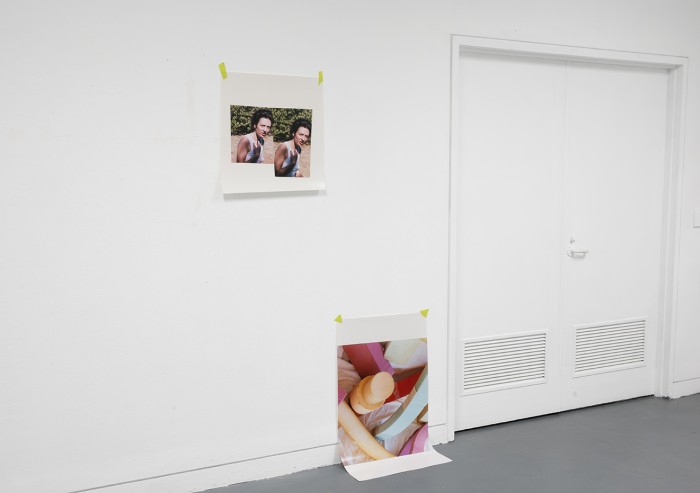 Photograph depicting two photographic prints of a persons face and a colourful sculpture. One is positioned higher up on the wall and the other lower to the floor touching the ground.