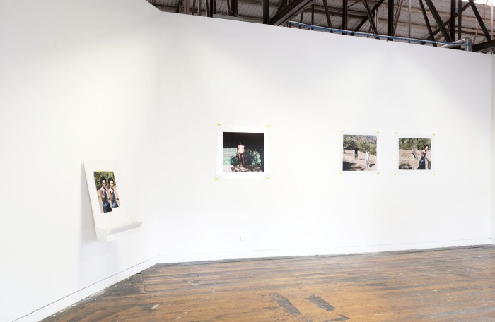Photograph depicting a plywood structure mounted off a wall with a photograph taped onto it and three other photographs on a joining wall depicting people standing in the sun