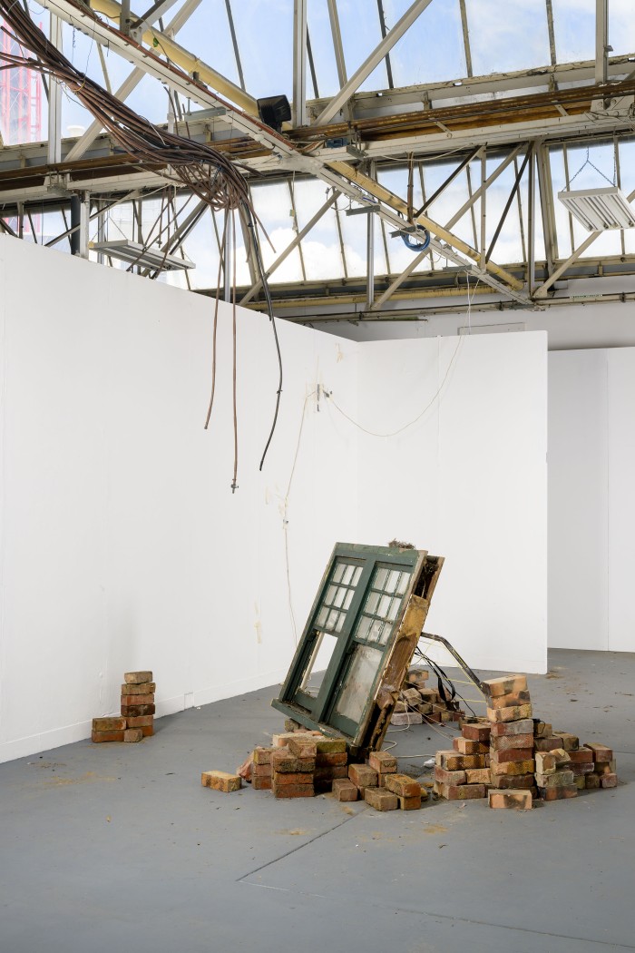 a green window disconnected from any wall is filled with spiderwebs and leans on piles of bricks and a clothes line in the middle of the gallery. there is a bundle of garden irrigation tubing hanging down from the ceiling skylights towards the window. white fluffy clouds and a blue sky are seen through rows of skylights.