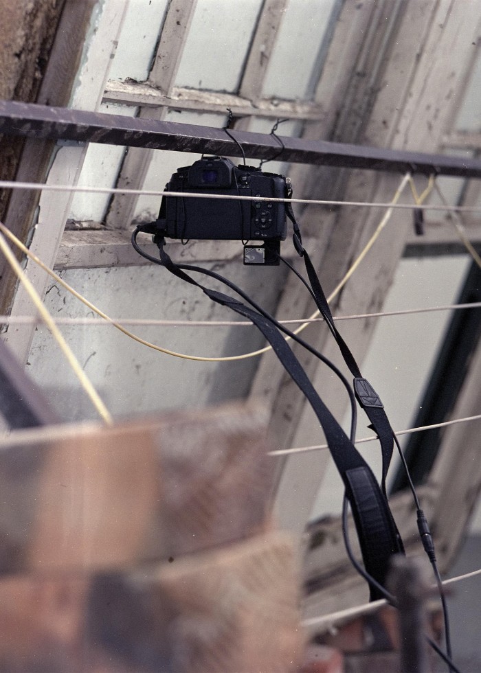 the side of the window painted white leans on a clothes line. Both objects are placed on the floor so that they are leaning on eachother and form a tent shape between them. a camera is tied to the clothes line and is pointing away from the viewer towards the web filled window. the camera cables drape over the clothes line strings.
