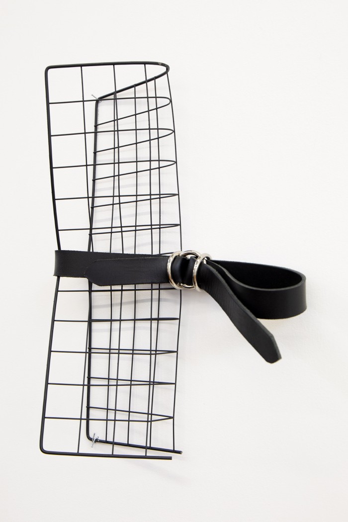 A photograph of a black metal grid folded lengthways in half, cinched by a length of leather woven through 3 chrome o-rings. It is hung on a white wall.  