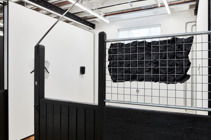 A photograph of an exhibition space bordered by a wooden fence painted black and a silver metal grid partition. There is a panel of leather pushing through the spaces of the grid from behind. Beyond the partition is are white walls, with two black metal and leather artworks hanging.