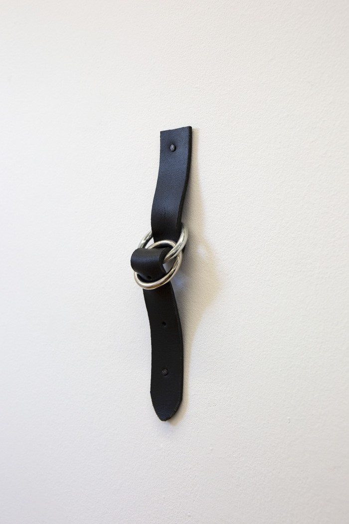 A photograph of a short length of black belt leather nailed to a white wall. The leather is woven through two chrome o-rings and bulging outwards slightly.