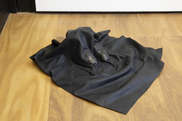 A photograph of a black leather shroud over a pair of boots, sitting on a wooden floor. The toes are polished to a shine, contrasting with the matte finish of the rest of the leather.