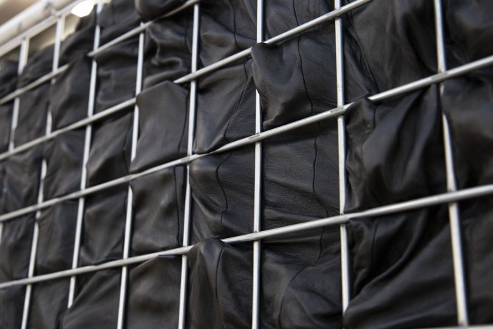 An photograph of black leather behind a silver metal grid. The leather is pushing through the spaces, there are impressions of fingers grasping out at the grid.