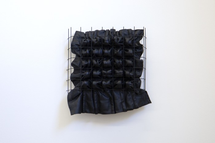 A photograph of a black metal grid embedded at its sides into a white wall. There is a panel of black leather pushing out from behind the grid, with bulky seams creating a pattern of lines and twists.  
