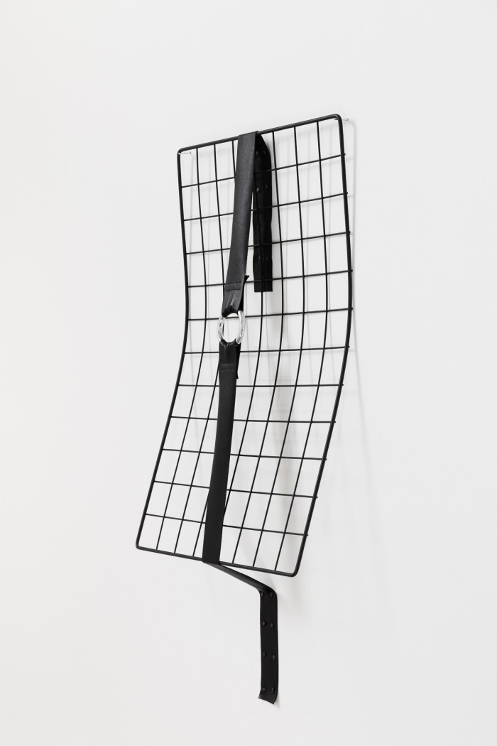 A photograph of a black metal grid againt a white wall. The grid is bent forward out from the wall, and is held in place by a vertical black leather strap with a chrome o-ring in the middle.