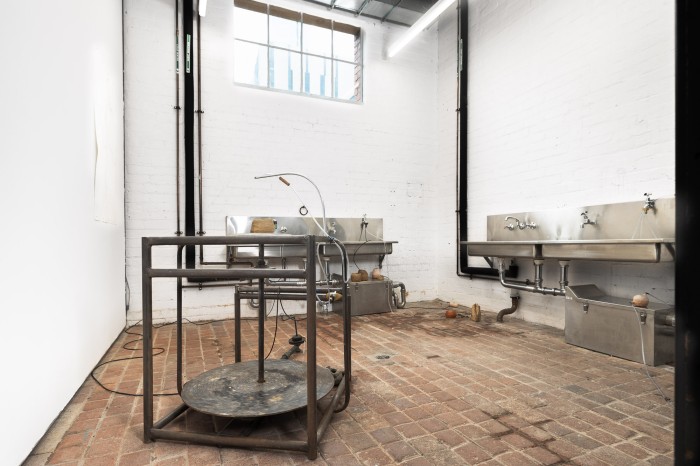 A small room with brick floor and two shiny metal sinks is filled with small clay vessels and a large steel spinning wheel structure. There are small black and clear tubes creating a system that moves water between the objects, leaking and being absorbed by the clay throughout the space.
