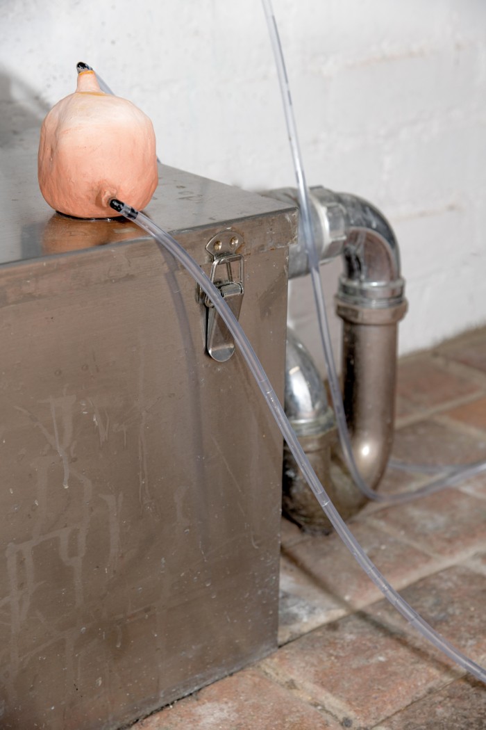 a small bisque-fired clay vessel leaks water that is fed into it thorugh clear pvc tubing. It sits amidst stainless steel plumbing.