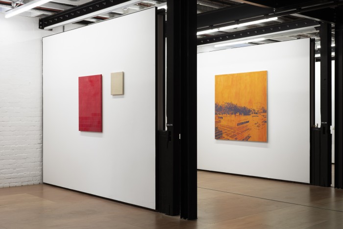 Installation view of portrait red and small beige painting on wall with big orange with grey landscape painting on the wall behind. The walls are parallel to each other.