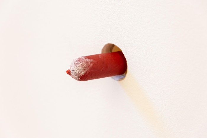 A close up image of the end of a red cocktail frankfurt protruding from a glory hole cut into the white plaster wall. A white powdered substance encrusts the end of the frankfurt.