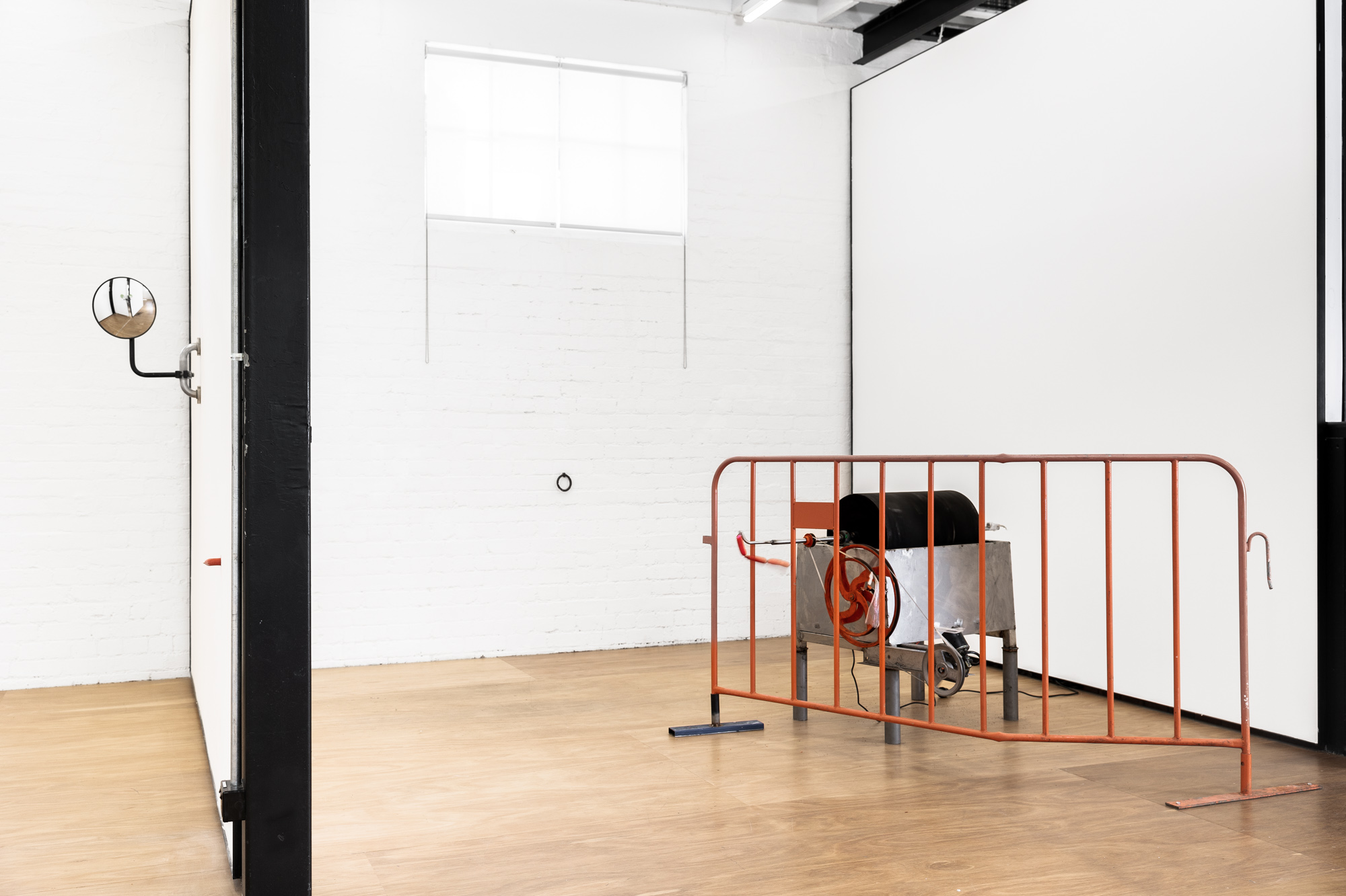 on the left side of the image is a dividing wall. There is a cocktail frankfurt potruding from this wall out of a glory-hole. Behind this, further into the space, is a black surveillance mirror mounted onto a piece of steel safety railing about 1.6 meters from the ground. The right side of the image looks into the second space on the other side of the dividimg wall. in this space there is a kinetic sculpture with a black spinning barrel. A piece of orange crowd-control fencing sits infront of it.