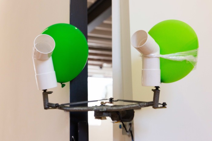 A windcreen wiper motor retaining part of its mechanical armature, is mounted to the intersection of two walls. two white L-shaped  poly pipes are mounted to each side. On the left side a green balloon is wedged between the polypip and the wall. On the right side a green ballon is attached to the poly pip with plastic wrap. the balloons and the pipes loosely resemble the clown game found at carnivals, as well as the way they slowly turn towards and away from each other.