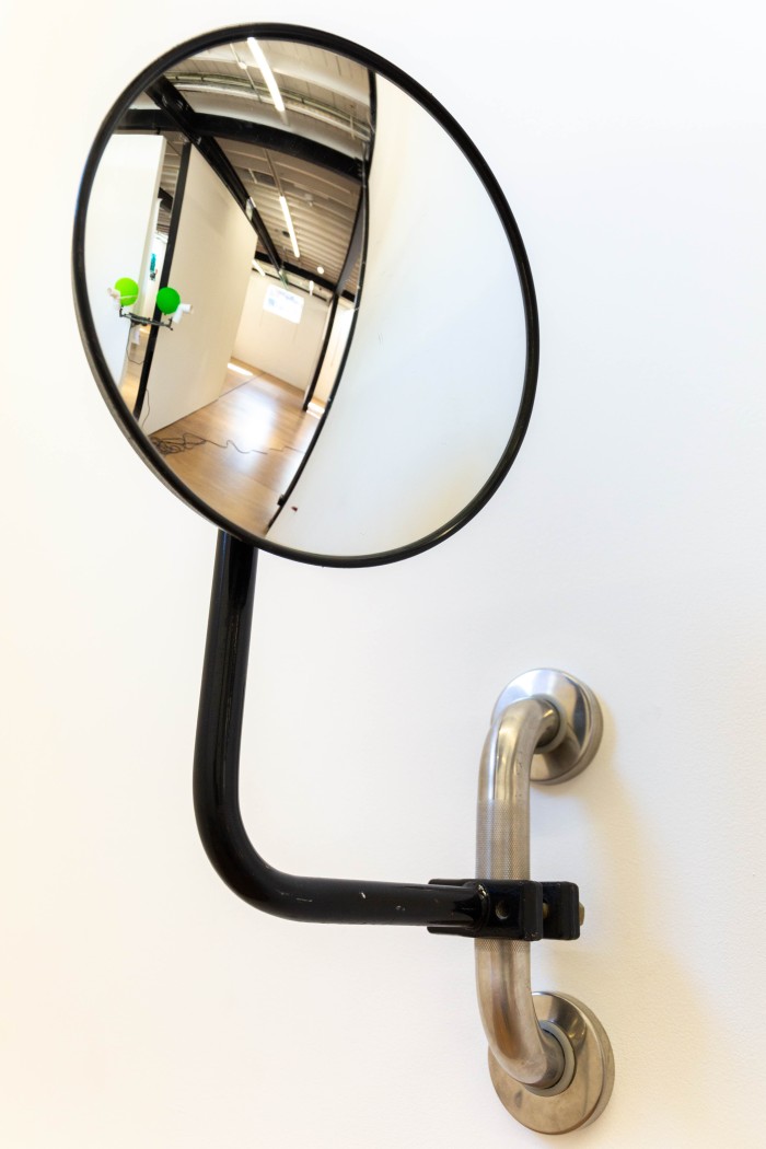 A close up image of a black convex surveillance mirror, mounted onto a piece of steel safety railing. In the mirror's reflection is the green balloon/polypipe sculpture and the end of the cocktail franfurt protruding from the glory-hole.