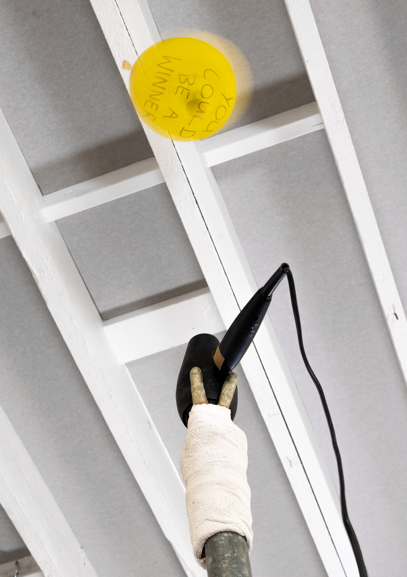 A golden pewter cast hand wrapped in a plaster bandage cast in a double finger "up yours" position. It simultaneously holds a black hairdryer. A yellow balloon hovers in the air above it. The words "YOU COULD BE A WINNER" are hand scrawled onto it with black texta.
