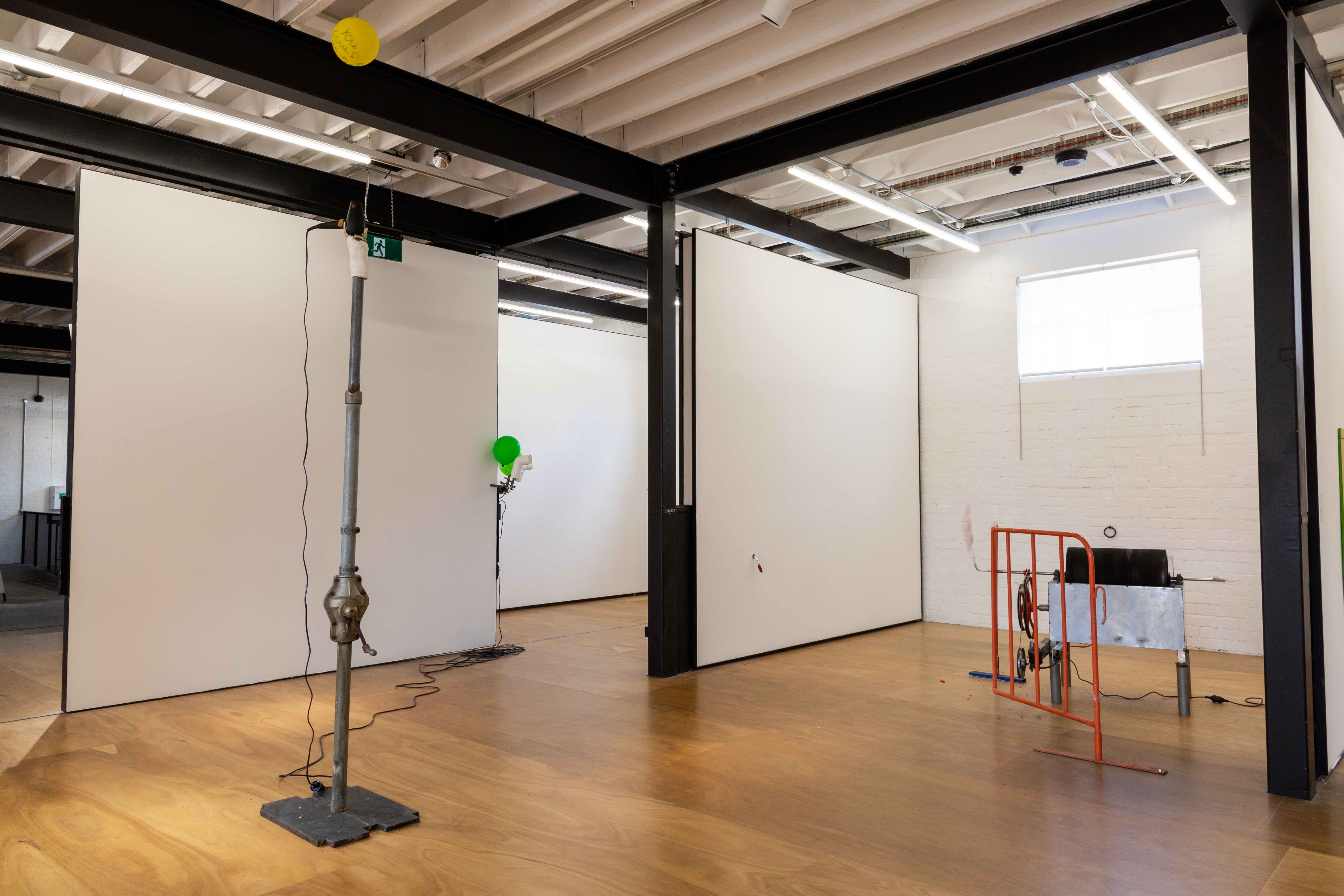 on the left side, in the foreground, is the central pole of a hills hoist. it acts as a plinth for the bandaged pewter hand, which is holding the hairdryer. the yellow balloon hovers in the airflow above, just below the ceiling. In the background, and centre of the image, is a green balloon wedged between an L-shaped polypipe and the outer corner of 2 walls. a dividing wall sits a few meters infront of this, and a piece of bend steel with a cocktail frankfurt shoved onto it's end is protruding from this wall, approximately 1 metre up from the floor. To the right of the image is a bent piece of orange crowd control fencing encasing a large kinetic sculpture with a black spinning barrel.