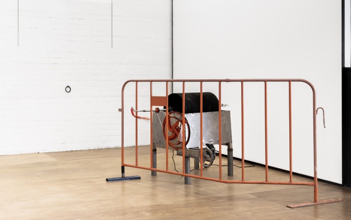 a stainless steel metal box on 4 legs encases the bottom half of a charcoal covered barrel. The barrel spins at high speed via a 12 v motor and a system of orange pulleys connected via plastic-wrap belts. a piece of orange crowd-control fencing is bent arount to partially cover the pulleys. Steel poles extend from both ends of the barrel and spin with it. the far end has a decaying parsnip attached to it via plastic wrsap. The closest end extends through the crowd control fencing and has a string of frankfurts attached to it. the spinning of the machine propells the frankfurts into a dance.