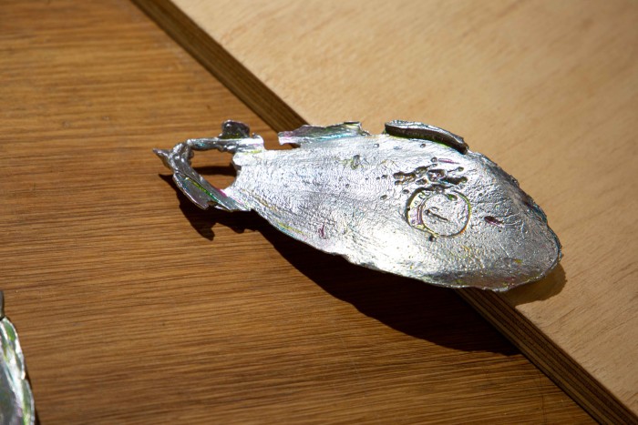A silver shiny cuttlebone is resting on the edge of a raw plywood structure that looks like it could have been a ramp. It has an impression of a ring at its centre.