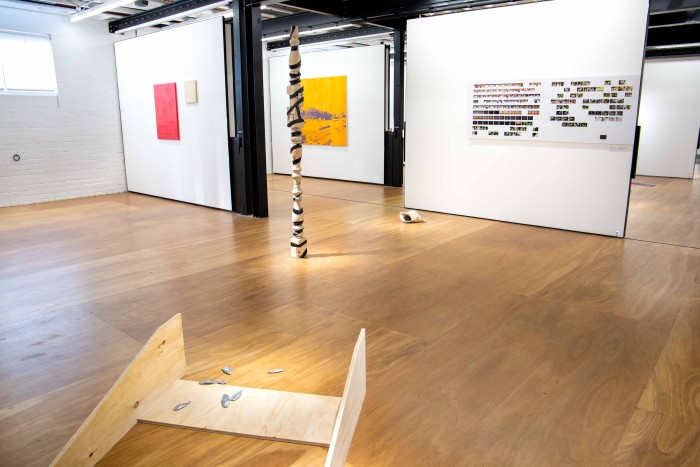 A wooden ramp-like structure with aluminium cast cuttlefish leads out into an open installation space, with a vertical sculpture of three sandblasted table legs stacked atop eachother, and a grid of photographs.