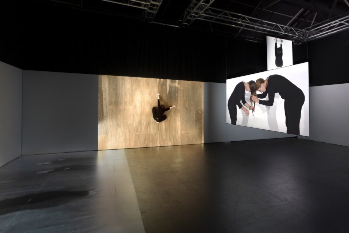 Three projections of people on a wall and screens, one is of a woman on a wooden floor and one is of two people plaiting their hair together. The third projection can only partially be seen and is of a woman hanging upside down and swinging.