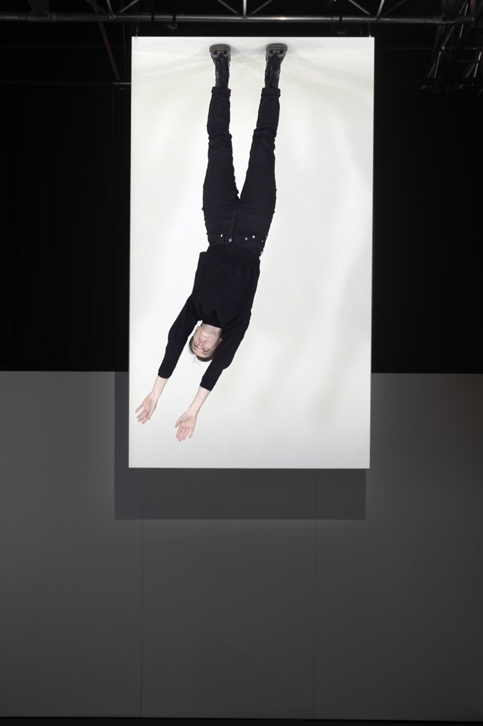 A person on a screen up high, hanging upside down with arms in the air. They look like they're swinging.