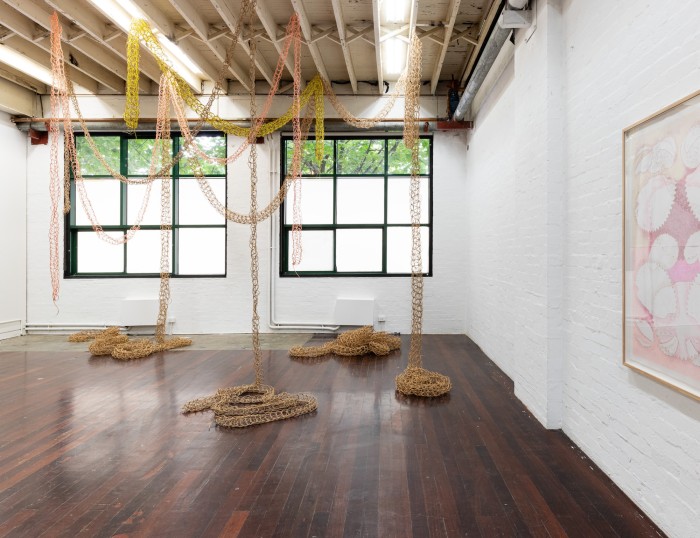 A room in which a series of loosely knitted tubes in brown, pink and fluorescent yellow are suspended from the roof. A cropped drawing sits to the right of the frame.