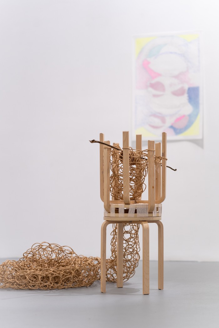 A pair of upturned wooden stools are taped together to create a loom. A rope of twisted brown craft paper is knitted through the legs of the stools and drapes in coils to the floor. In the background is an out of focus drawing on paper in fluorescent colours.