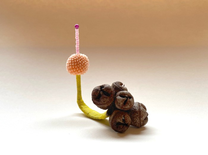 An assemblage in which a dressmakers pin threaded with soft pink beads pokes out the top of a small peach coloured ball. The ball sits atop the stem of a cluster of native seedpods which has been wrapped in bright yellow tape.