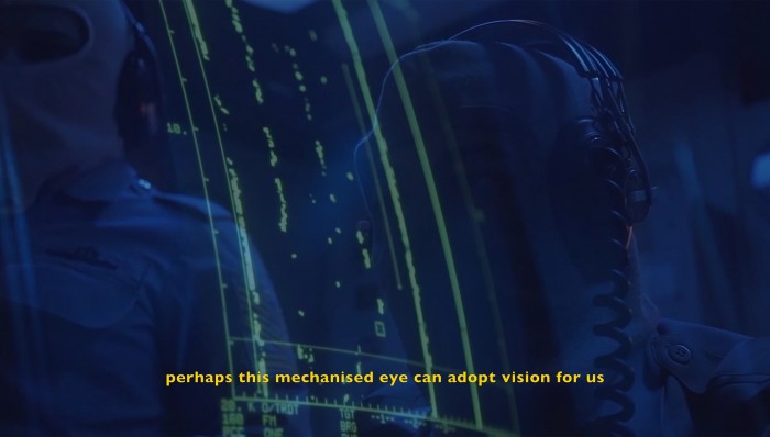 Two engineers sit in a submarine looking at a sonar interface, the neon sonar markers on the screen are overlaid on their faces like a reflection.