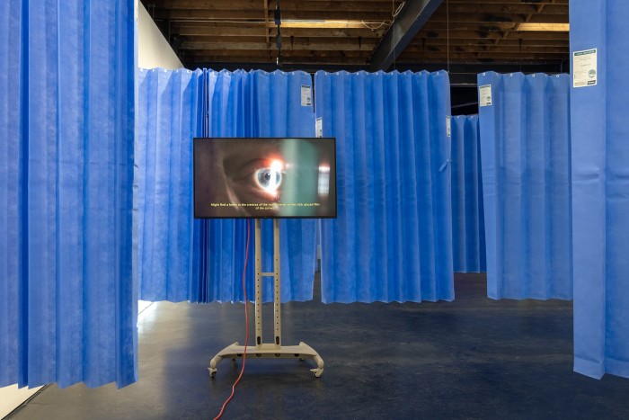A large beige TV stand holds a LCD monitor screen playing a video depicting a close-up view of an eye. Surrounding the stand are hanging blue disposable hospital curtains.