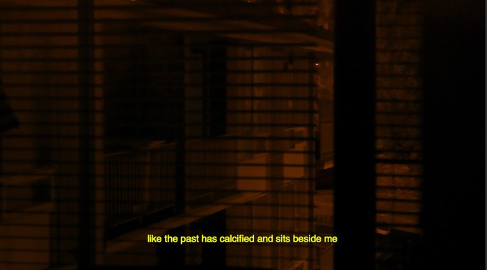 A dimly lit view through a window with venetian blinds. Orange streelight seeps into the room. A yellow subtitle reads "like the past has calcified and sits beside me"