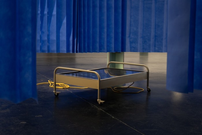 A large beige metal trolley holds a LCD monitor screen playing a video depicting the facade of a hospital. Surrounding the trolley are hanging blue disposable hospital curtains and a round mint green ply wood stool.