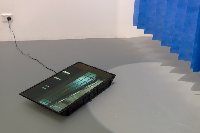 A TV monitor lies flat on the ground, on the screen is a scene of a hospital facade at night lit by green streetlights. The TV is overlaid with a round spotlight. Next to the TV hangs a row of disposable hospital curtains. The edge of the curtains bisects the spotlight creating a zigzag shadow.