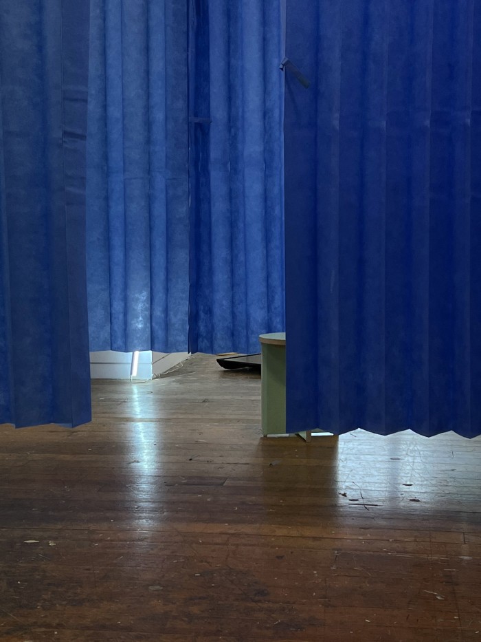 In a room with a wooden floor hang rows of blue disposable hospital curtains. On the ground lie several LED lights. Between the curtains sits a mint green plywood stool.